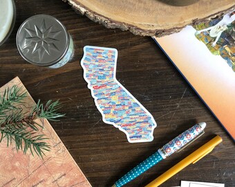 May include: A sticker of the state of California with the names of cities and towns printed in a colorful pattern on a wooden surface.