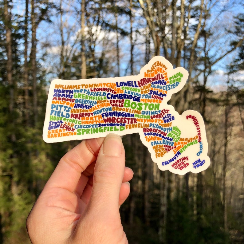 May include: A sticker shaped like the state of Massachusetts with the names of cities and towns in the state written inside the outline. The sticker is white with colorful text. The sticker is being held by a hand against a blurred background of trees.