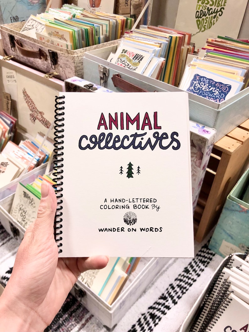 Puede incluir: Un libro para colorear escrito a mano titulado "Animal Collectives" se sostiene en una mano. La portada presenta el t&iacute;tulo en escritura roja y azul, con una peque&ntilde;a ilustraci&oacute;n de &aacute;rboles. El libro est&aacute; encuadernado en espiral y rodeado de otros art&iacute;culos de papeler&iacute;a.