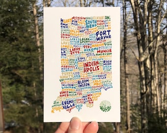 May include: A postcard featuring a colorful map of Indiana, with city names forming the state's outline. The text "Wander on Words" is printed in a green circle on the bottom right corner. The card is held against a backdrop of trees.