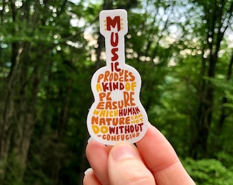 Pode incluir: Um adesivo branco em forma de guitarra com o texto "A música produz um tipo de prazer do qual a natureza humana não pode prescindir -Confúcio" em letras marrons, laranja e amarelas.