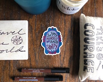May include: A sticker with a mirror frame design and the text "Loving yourself isn't vanity it's sanity". The sticker is on a wooden surface with other items.