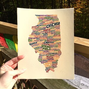 May include: A print of the state of Illinois, filled with the names of cities in various colors. The word "Chicago" is prominently displayed. The artwork is held by a hand, with a natural outdoor background.