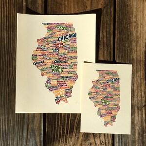 May include: Two art prints of the state of Illinois, filled with city names in various colors. The prints are on cream-colored paper and are displayed on a wooden surface. The larger print is approximately 11x14 inches, and the smaller one is about 5x7 inches.