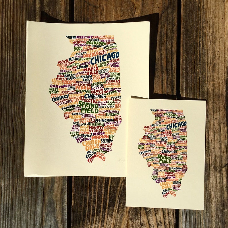 May include: Two art prints of the state of Illinois, filled with city names in various colors. The prints are on cream-colored paper and are displayed on a wooden surface. The larger print is approximately 11x14 inches, and the smaller one is about 5x7 inches.
