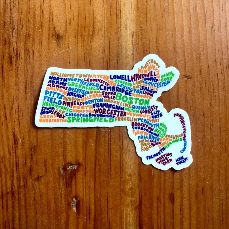 May include: A colorful sticker shaped like the state of Massachusetts with the names of cities and towns in the state written inside the outline. The sticker is on a wooden surface.