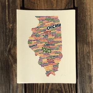 May include: A print of the state of Illinois, filled with the names of cities in various colors. The cities are arranged to form the shape of the state. The background is a wooden surface.