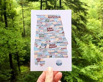 Può includere: Un biglietto bianco con un contorno blu, rosso e giallo dello stato dell'Alabama. Il contorno è riempito con i nomi delle città e dei paesi dell'Alabama. Il testo "Wanderlust of Words" è in fondo al biglietto.