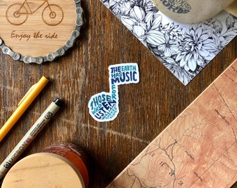 Pode incluir: Um adesivo azul e branco com o texto "The Earth Has Music For Those Who Listen" disposto em forma de nota musical. O adesivo está sobre uma superfície de madeira.