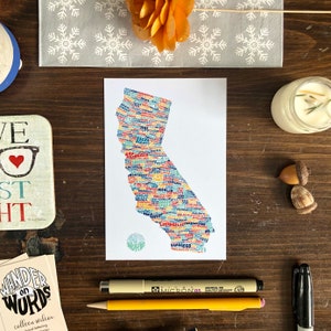 May include: A white postcard with a colorful map of California made of words. The map is outlined in blue and filled with different colored words, including city names. The words "Wander on Words" are in a circle in the lower left corner of the map.
