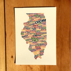 May include: A white rectangular print featuring the state of Illinois, filled with the names of cities in various colors. The word "Chicago" is prominently displayed. The print is on a wooden surface.