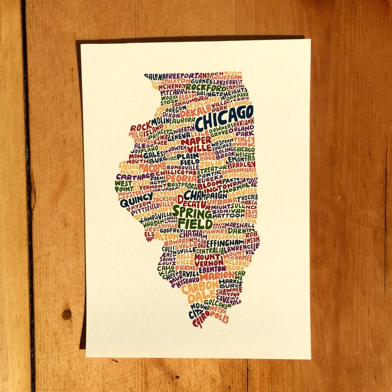 May include: A white rectangular print featuring the state of Illinois, filled with the names of cities in various colors. The word "Chicago" is prominently displayed. The print is on a wooden surface.
