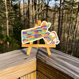 May include: A colorful sticker shaped like the state of Massachusetts with the names of cities and towns in the state written inside the outline. The sticker is on a small wooden easel.
