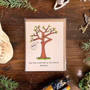 May include: A greeting card with a tree illustration and the text "How does a tree feel in the spring? Relieved." The tree is brown with green leaves and a speech bubble that says "Phew". The card is in a clear plastic sleeve.