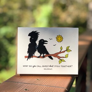 May include: A greeting card with a whimsical illustration of two black birds perched on a branch with a yellow sun. The card features the text: "What do you call birds that stick together? VELCROWS."