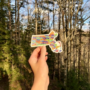 May include: A colorful sticker shaped like the state of Massachusetts with the names of cities and towns written inside the outline. The sticker is being held in a hand against a blurred background of trees.