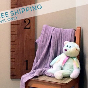 May include: A wooden growth chart with black numbers 1 and 2, mounted on a wall. A purple blanket and a light blue teddy bear are on a wooden chair in front of the chart.