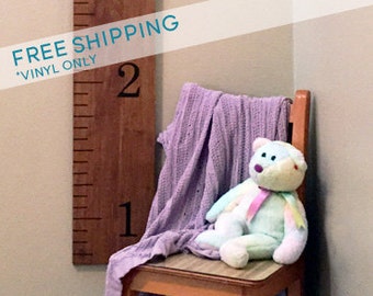 May include: A wooden growth chart with black numbers 1 and 2, mounted on a wall. A purple blanket and a light blue teddy bear are on a wooden chair in front of the chart.