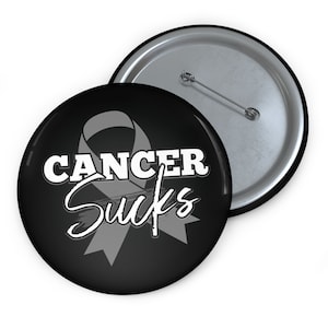 May include: A black button with a gray ribbon and the text "Cancer Sucks" in white. The button has a silver back.
