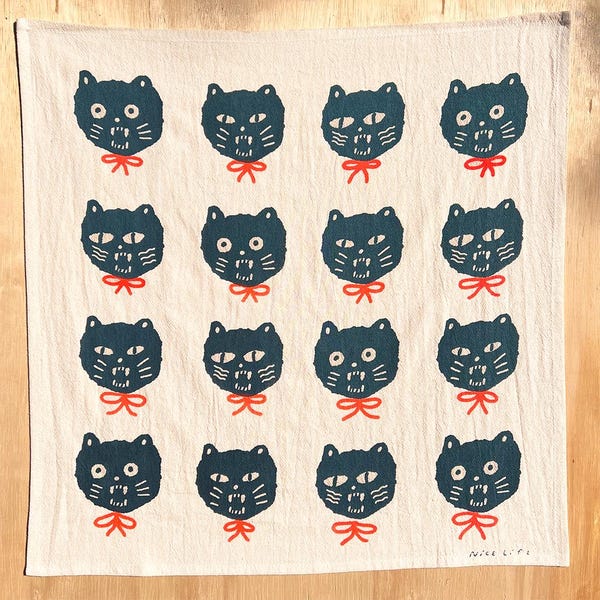 May include: A cream-colored kitchen towel featuring a repeating pattern of stylized black cat faces with red bow ties. The towel has a simple, minimalist design, with the words "Nice Life" printed in the bottom right corner.