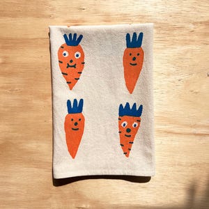 May include: A cream-colored kitchen towel featuring four cartoon carrots with orange bodies and blue tops. The carrots have simple facial features, including eyes and smiles, and are arranged in a 2x2 grid.