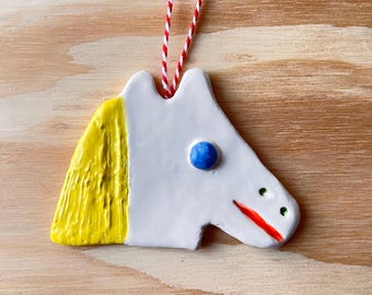 May include: White ceramic ornament shaped like a horse's head with yellow hair, a blue eye, and a red mouth.