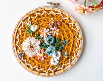 May include: A round embroidery hoop with a mustard yellow wool background and a floral embroidery design featuring pink, white, and blue flowers.
