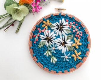 May include: A blue embroidery hoop with white flowers and yellow accents. The flowers are embroidered with white yarn and have black centers. The hoop is surrounded by a bouquet of colorful flowers.