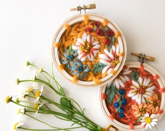 May include: Two small embroidery hoops with floral designs. The hoop on the left has a yellow background with white, orange, red, and blue flowers. The hoop on the right has an orange background with white, yellow, and blue flowers. A bouquet of white daisies with green stems is tied with a yellow ribbon.