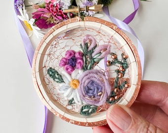 May include: A small embroidery hoop with a floral design. The flowers are made of purple, white, and green yarn and are surrounded by copper wire. The hoop is made of wood and has a copper finish.