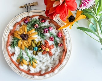 May include: A white embroidery hoop with a floral design. The design features yellow, orange, red, green, and blue embroidery floss. The hoop is surrounded by a brown wooden frame.