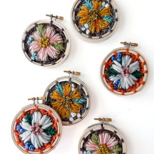 May include: A collection of six embroidered floral designs in wooden hoops. The flowers feature various colors, including pink, orange, yellow, blue, and green, with detailed stitching. The hoops are arranged on a white background, showcasing the handcrafted embroidery.