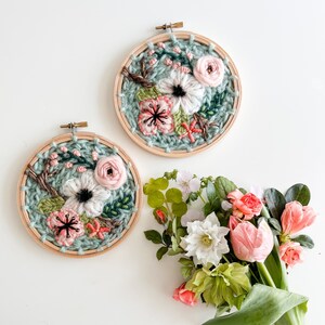 May include: Two embroidery hoops with floral designs. The hoops are made of wood and have a light green background. The flowers are pink, white, and green. A bouquet of pink and white flowers sits below the hoops.