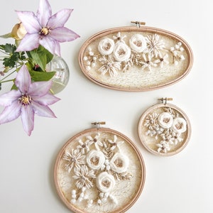 May include: Two embroidery hoops with white floral designs on a white background. The larger hoop is oval shaped and the smaller hoop is round. Both hoops have a wooden frame.