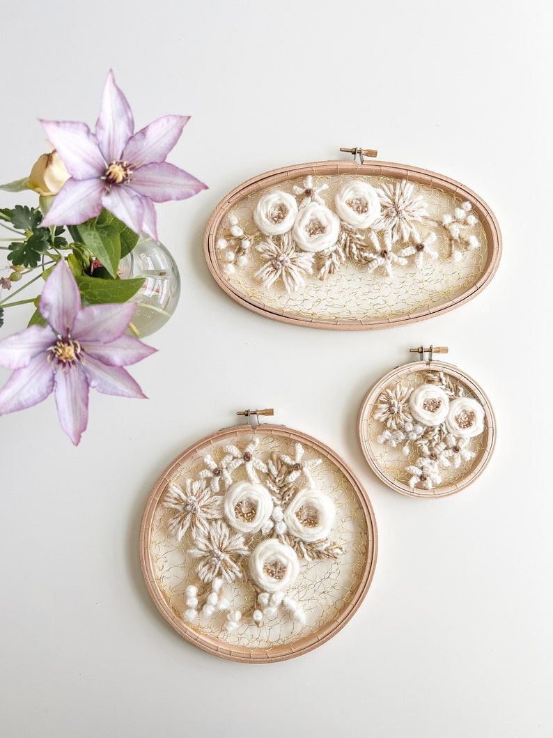 May include: Two embroidery hoops with white floral designs on a white background. The larger hoop is oval shaped and the smaller hoop is round. Both hoops have a wooden frame.