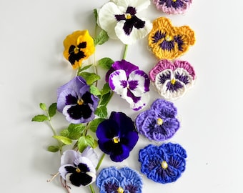 May include: A collection of crocheted and real pansies in various shades of purple, blue, yellow, and pink. The crocheted pansies are arranged in a circle around the real pansies.