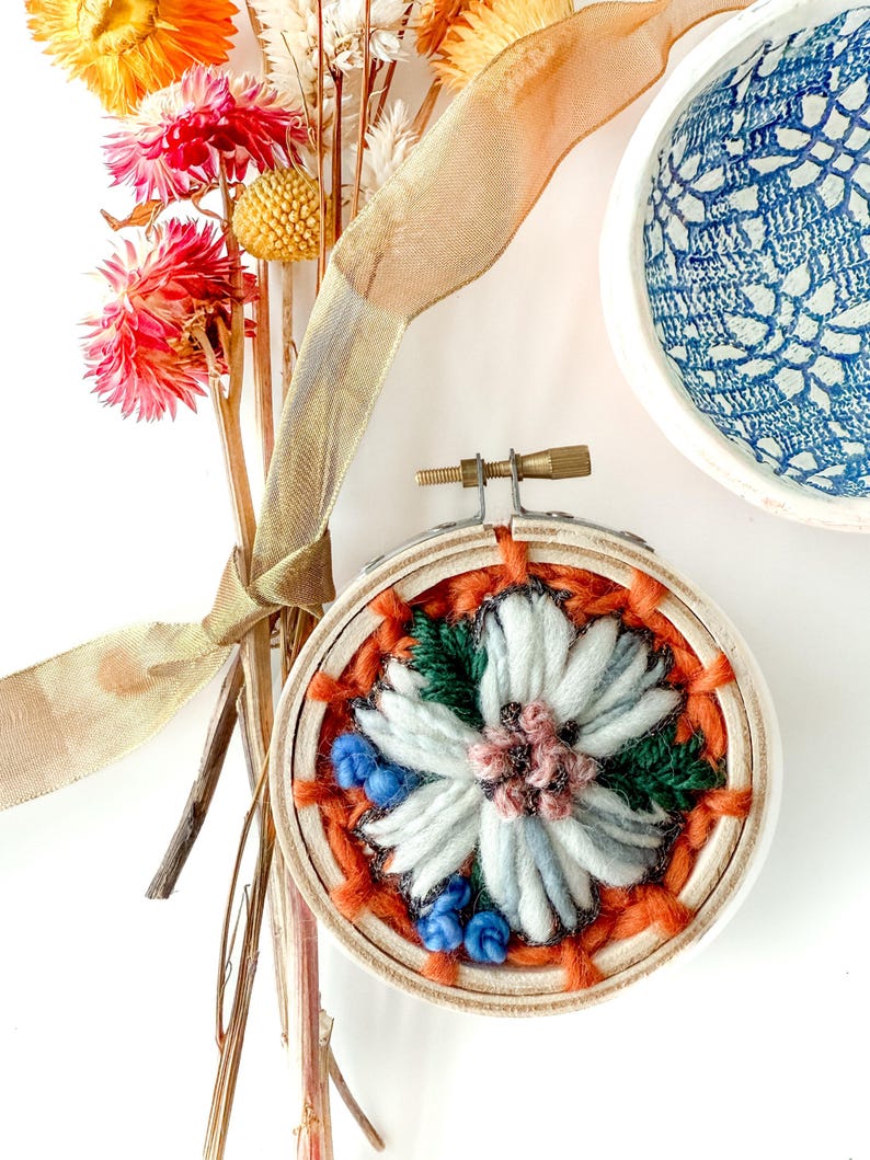 Pu&ograve; includere: Un telaio da ricamo fatto a mano con un fiore bianco, dettagli grigi e rosa e filo arancione. Il telaio &egrave; ambientato su uno sfondo di fiori secchi e una ciotola a motivi blu e bianchi. Un nastro dorato aggiunge un tocco decorativo.