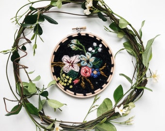 May include: A floral embroidery hoop art piece featuring a bouquet of colorful flowers on a black background. The embroidery is set in a wooden hoop and surrounded by a wreath of vines and white flowers, all set against a white background.