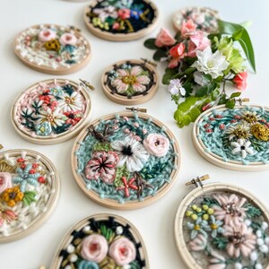 May include: A collection of nine embroidery hoops with floral designs. The hoops are made of wood and have a variety of colors, including pink, green, white, and black. The flowers are embroidered with yarn and beads.