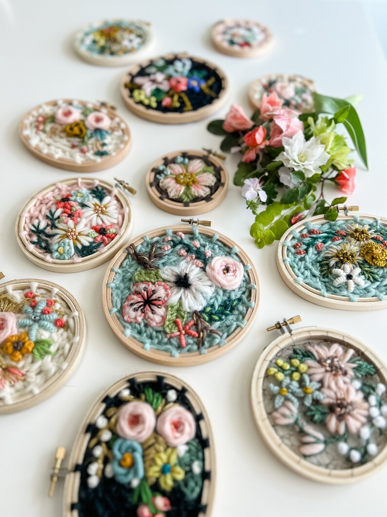 May include: A collection of nine embroidery hoops with floral designs. The hoops are made of wood and have a variety of colors, including pink, green, white, and black. The flowers are embroidered with yarn and beads.