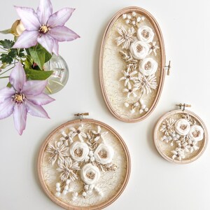May include: Three embroidery hoops with white floral designs on a white background. The hoops are different sizes and are all framed in natural wood.