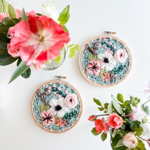 May include: Two embroidery hoops with floral designs. The hoops are made of wood and have a light blue background. The flowers are pink, white, and green.
