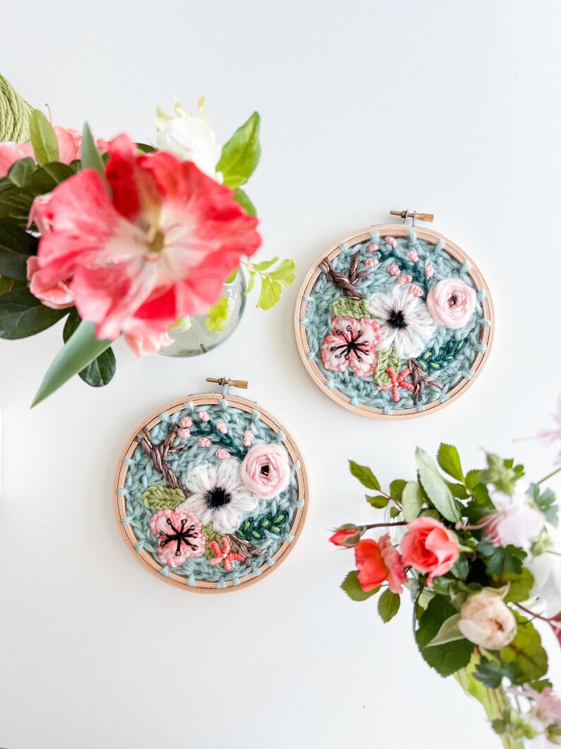 May include: Two embroidery hoops with floral designs. The hoops are made of wood and have a light blue background. The flowers are pink, white, and green.