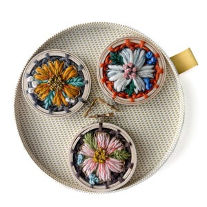 Pu&ograve; includere: Tre piccoli telai da ricamo rotondi con disegni floreali colorati. I telai sono posizionati su un vassoio con motivi oro e bianco. I fiori presentano sfumature di arancione, blu, rosa e verde, con cuciture e trame dettagliate.
