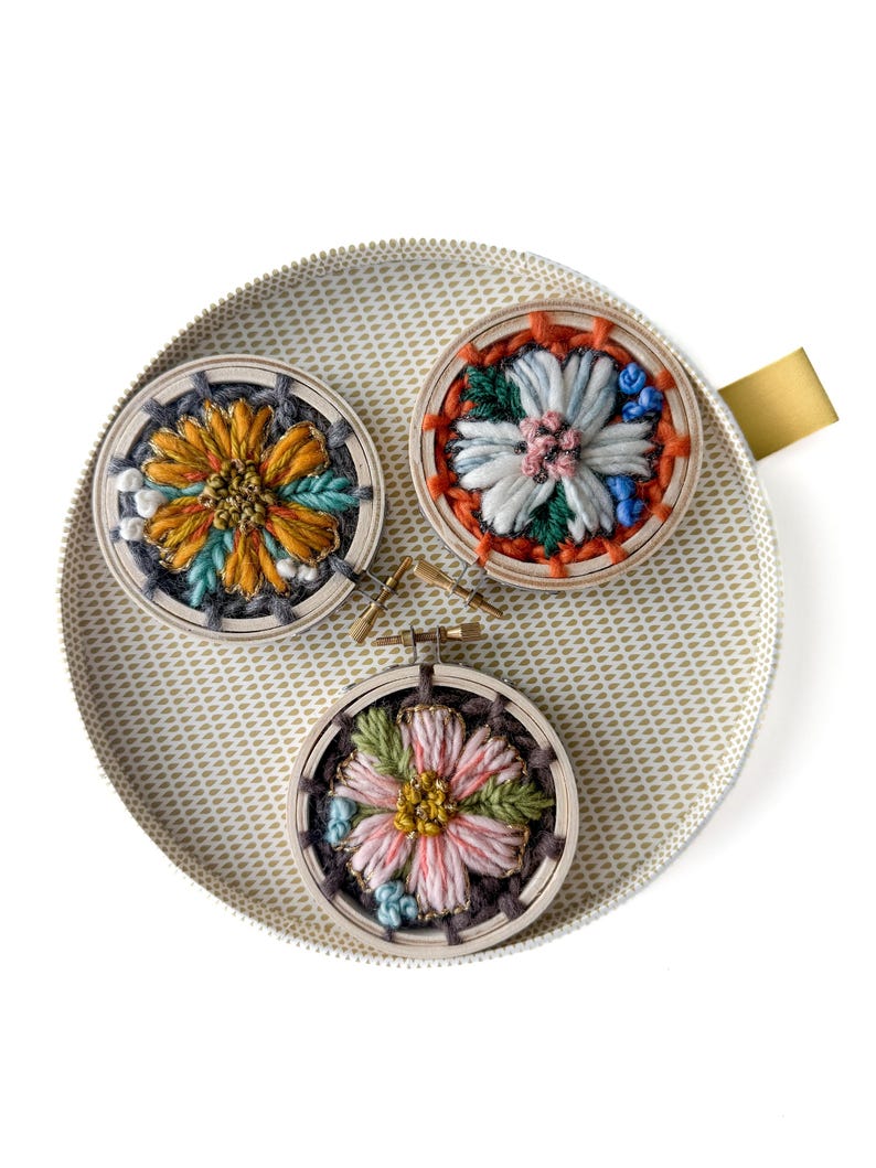Pu&ograve; includere: Tre piccoli telai da ricamo rotondi con disegni floreali colorati. I telai sono posizionati su un vassoio con motivi oro e bianco. I fiori presentano sfumature di arancione, blu, rosa e verde, con cuciture e trame dettagliate.