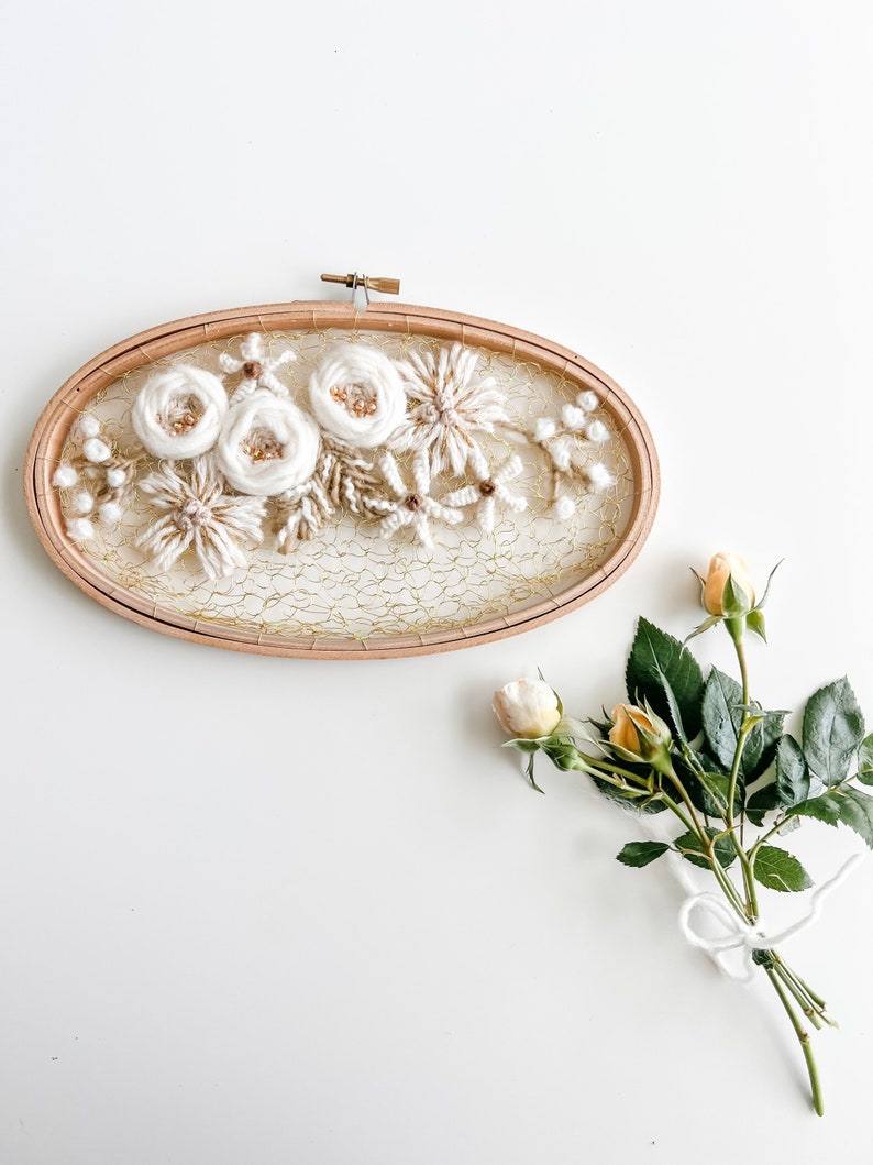May include: A white floral embroidery hoop art piece with a gold mesh background. The hoop is made of wood and has a simple design. The flowers are made of yarn and are arranged in a cluster. The hoop is hanging on a white wall.