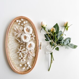 May include: A white and beige floral embroidery hoop art piece with a gold mesh background. The hoop is oval shaped and is surrounded by a wooden frame. A bouquet of yellow roses with green leaves is on the right side of the image.