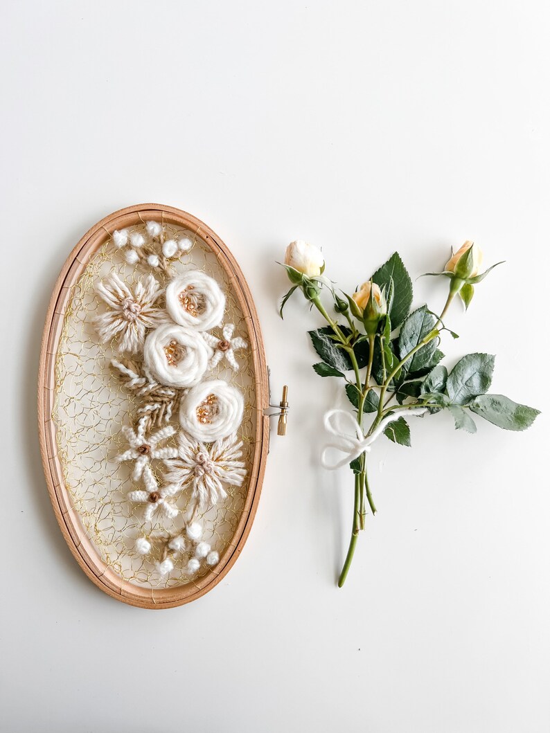 May include: A white and beige floral embroidery hoop art piece with a gold mesh background. The hoop is oval shaped and is surrounded by a wooden frame. A bouquet of yellow roses with green leaves is on the right side of the image.