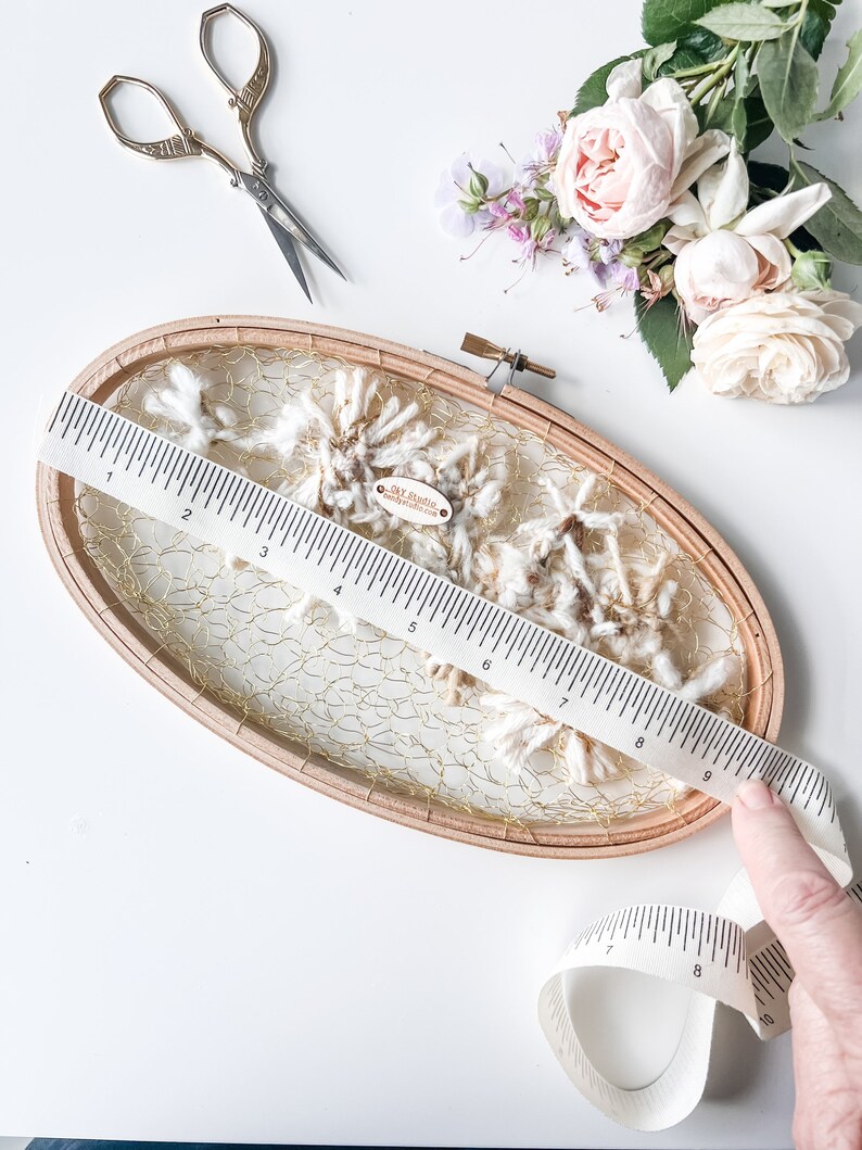 May include: A wooden embroidery hoop with a gold mesh and white yarn inside. A ruler and a measuring tape are also in the image. The tag on the yarn reads "Clay Studio Handmade with Love".