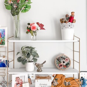 May include: A white shelf unit with gold metal accents, featuring a variety of decorative items, including a vase with pink and red flowers, a small potted plant, a bird figurine, a framed photo, a wooden box with the text "dream big", and a pair of embroidered gloves.